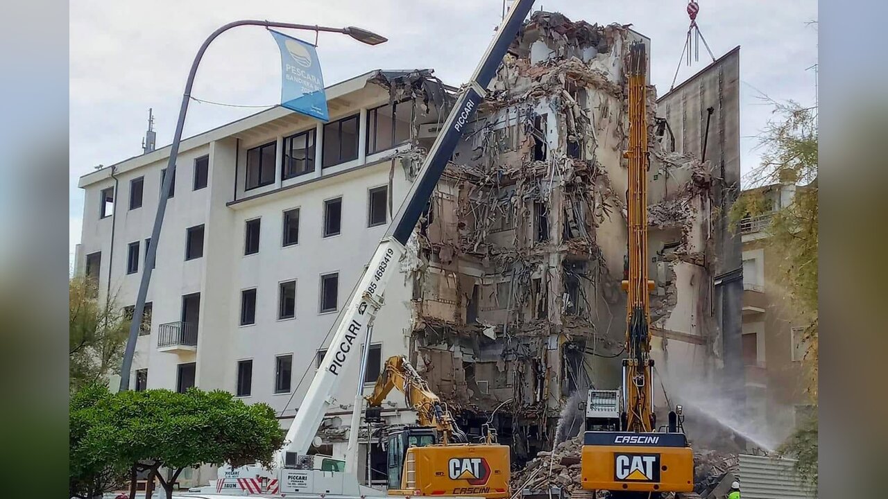 Demolizione di un edificio: escavatori e una gru davanti a un palazzo bianco per metà ridotto in macerie, con travi e solai a vista